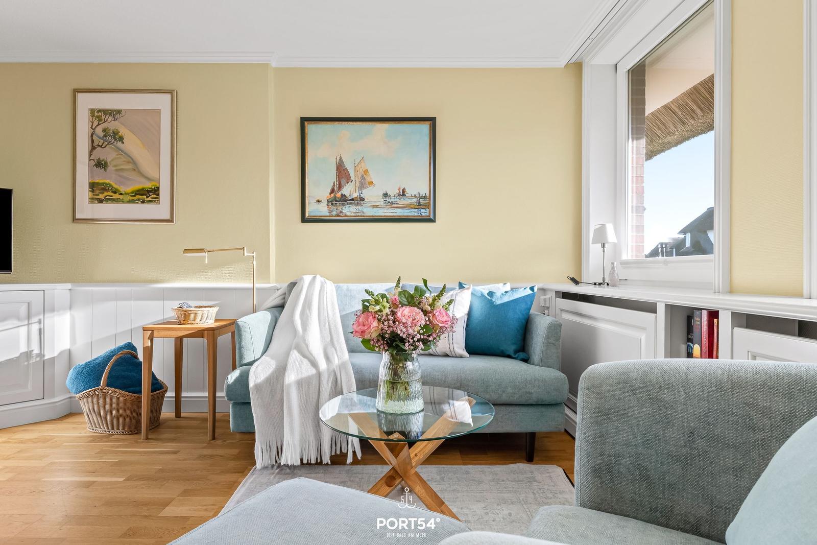Ein Wohnzimmer mit Sofa, Couchtisch und Wandbildern. Durchblick auf Außenbereich.