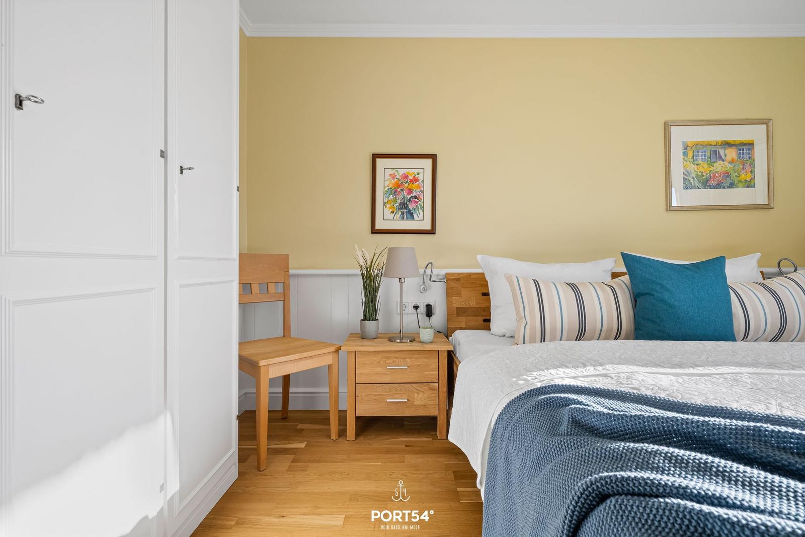 Schlafzimmer mit Bett, Nachttisch, Stuhl und zwei Bildern an der Wand.