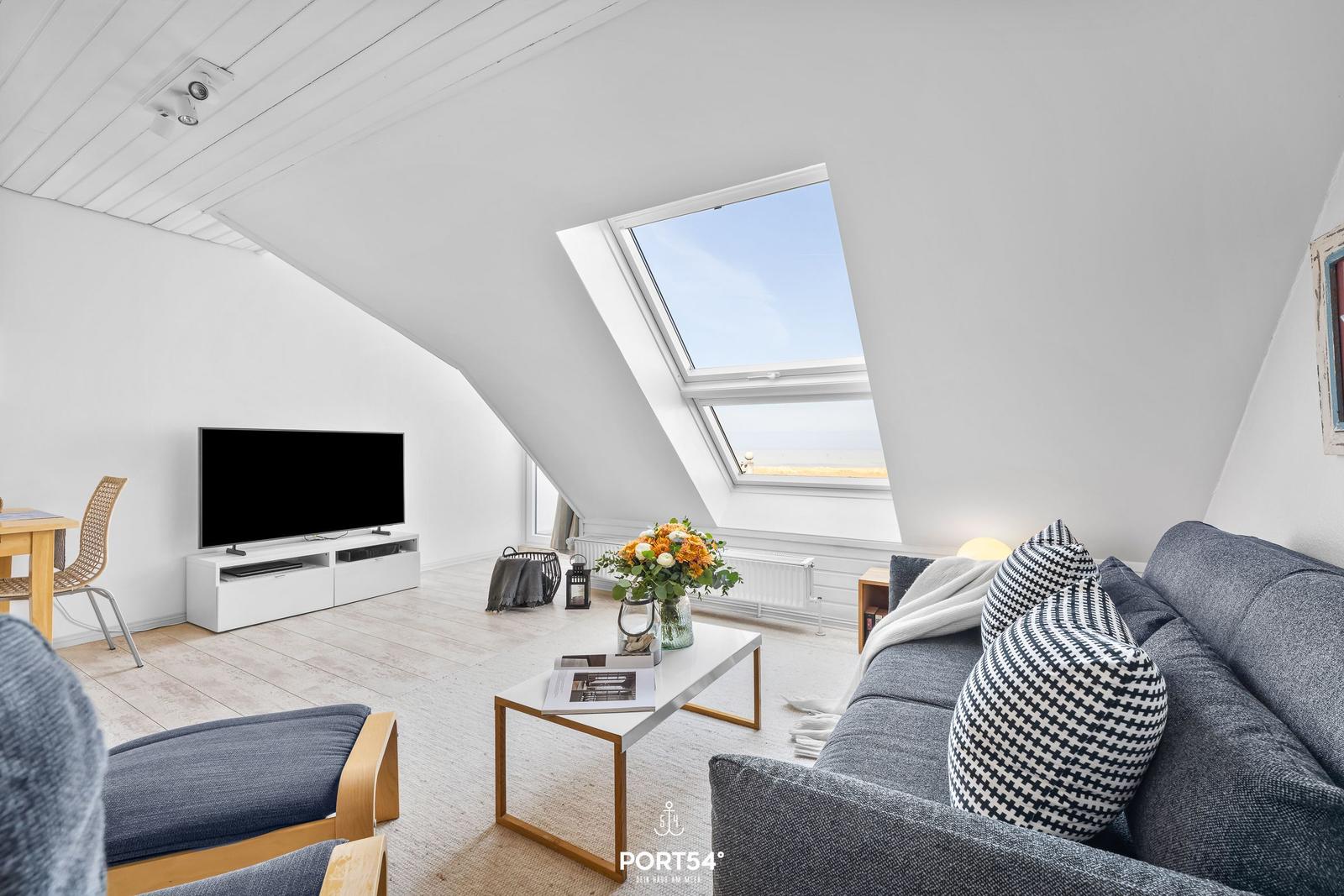 Wohnraum mit Dachfenster, Sofa, Fernseher und Tisch. Blick auf Strand.