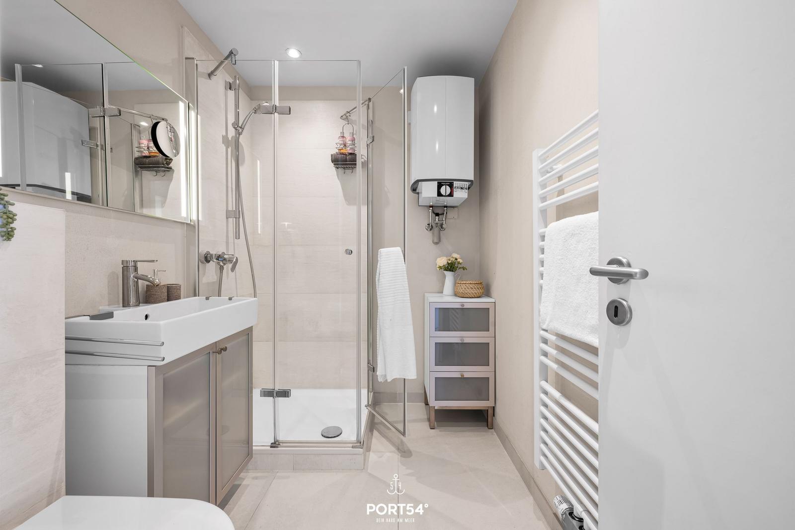 Badezimmer mit Dusche, Waschbecken und Heizung. Handtuchhalter und kleine Schränke sind sichtbar.