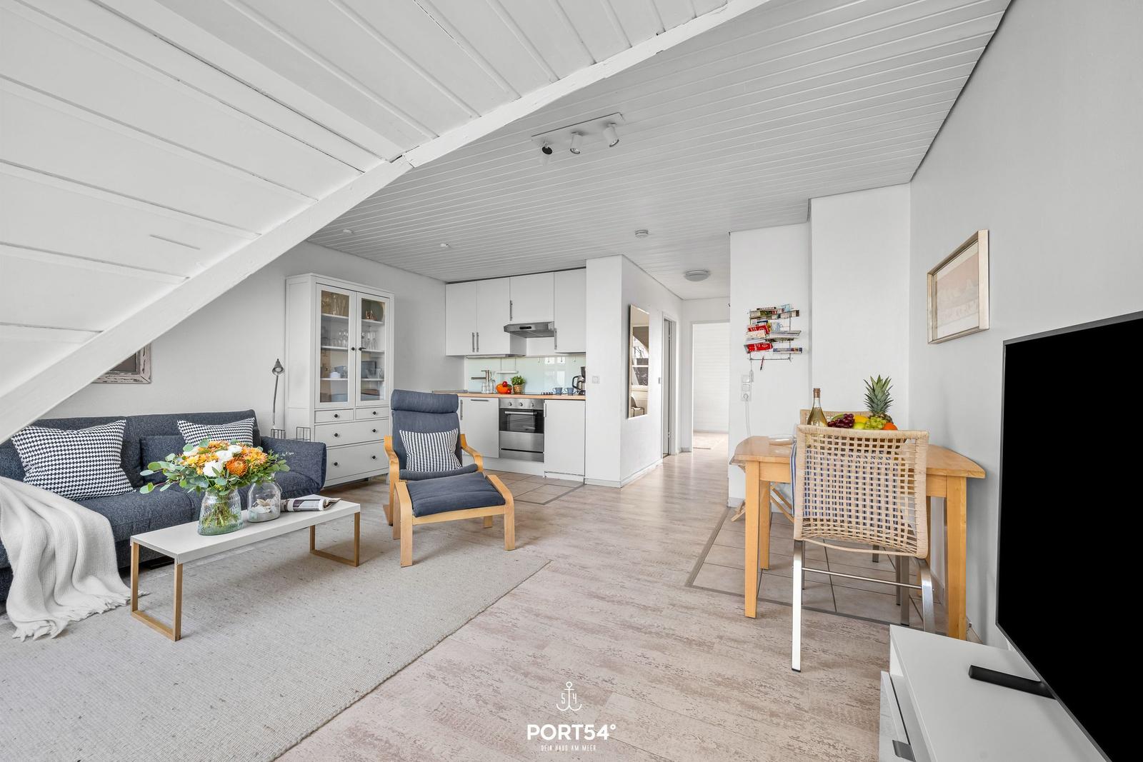Wohnbereich mit Sofa, Sessel und Küchenbereich. Holzboden und weiße Decke.