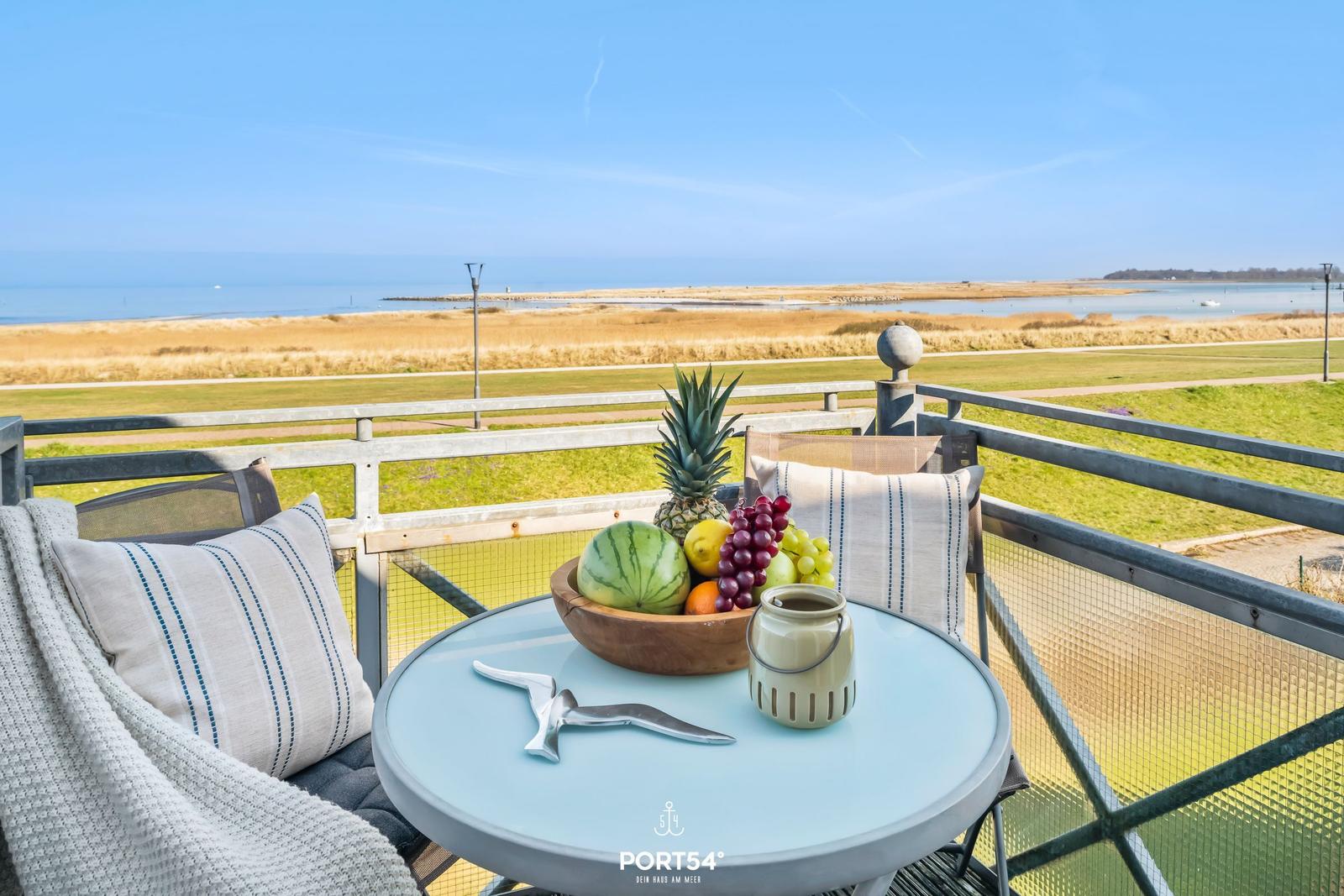Terrasse mit Tisch, Stühlen und Obstschale. Blick auf Grünfläche und Meer.