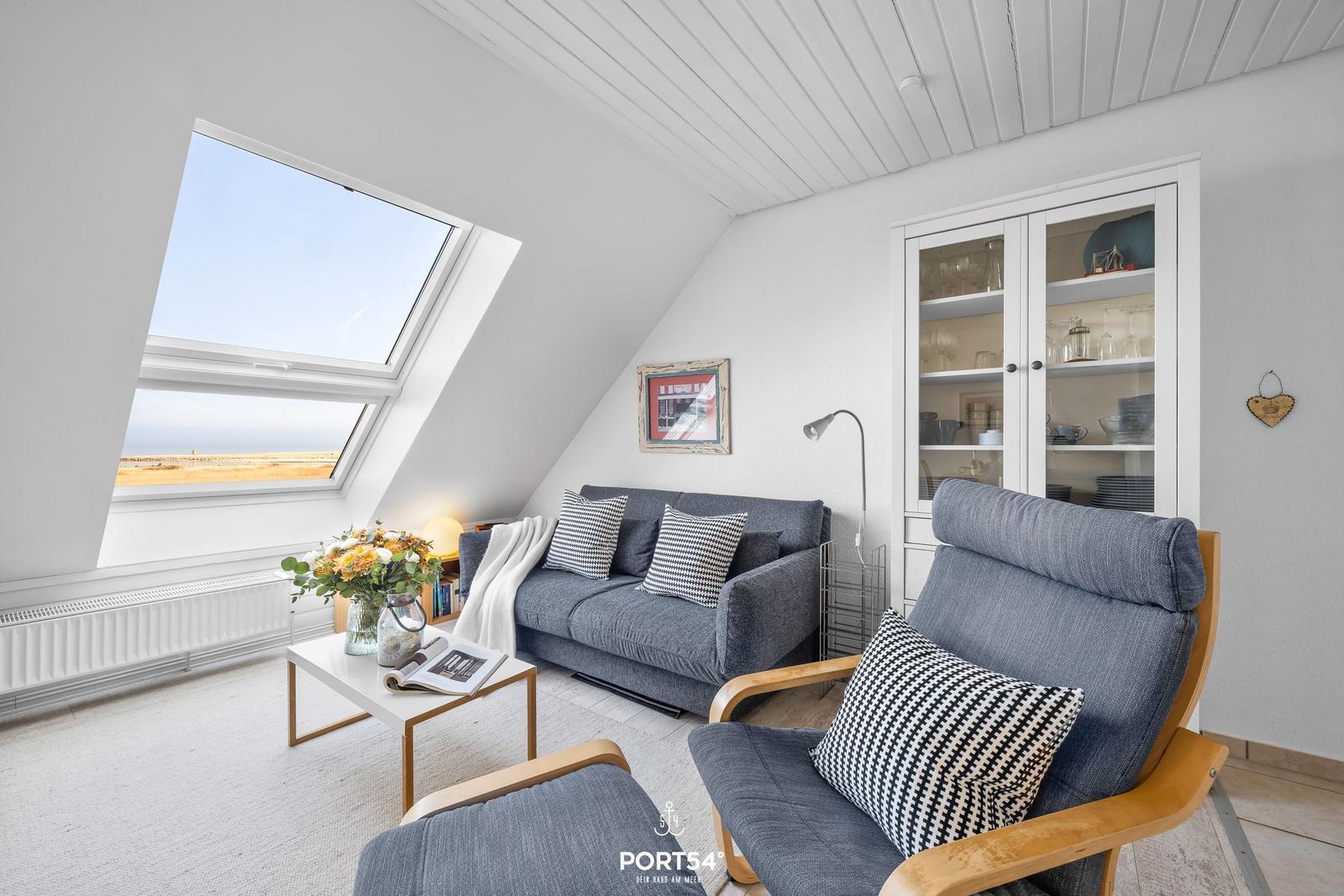 Dachgeschosswohnung mit großem Dachfenster, Sofa und Sessel. Blick auf Küstenlandschaft.