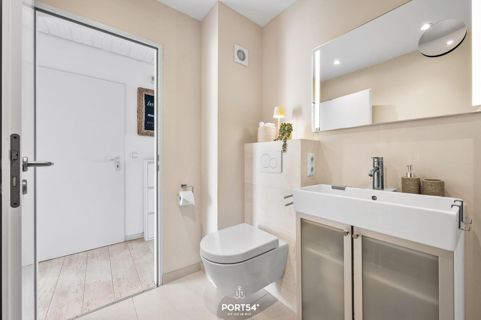 Badezimmer mit Toilette, Waschbecken und Spiegel. Tür führt in einen Flur.
