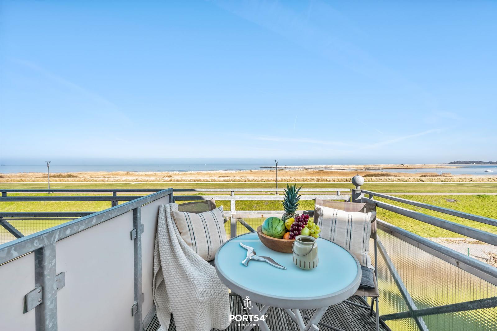 Balcony with table, chairs, and fruit bowl overlooking a grassy area and sea.