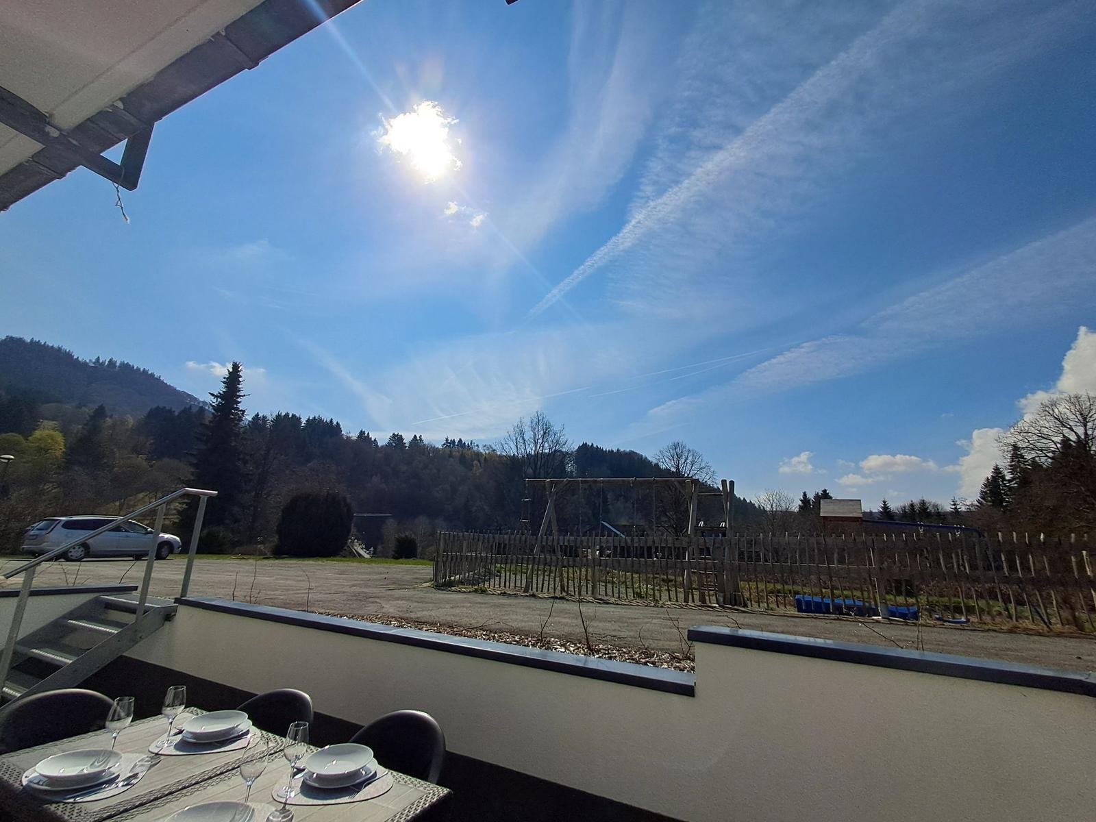 Terrasse mit Tisch und Stühlen. Blick auf Wald und Himmel mit Sonne.