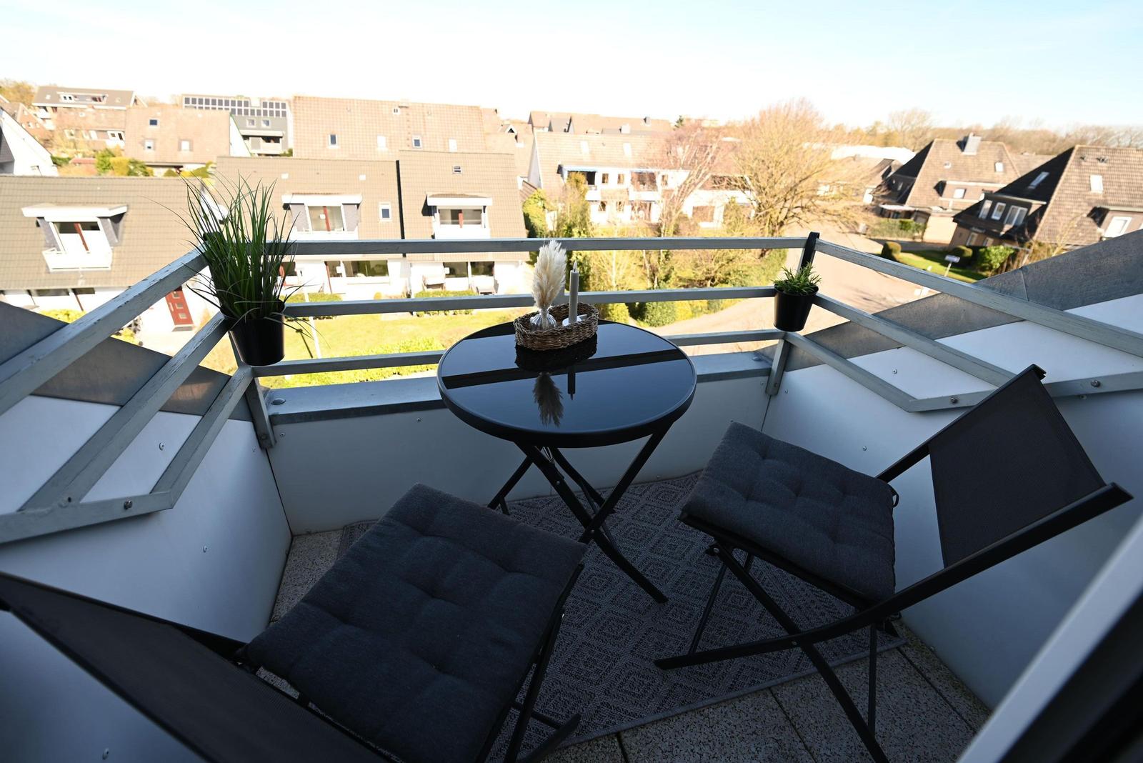 Balcony with table, two chairs, and potted plants overlooking residential area.