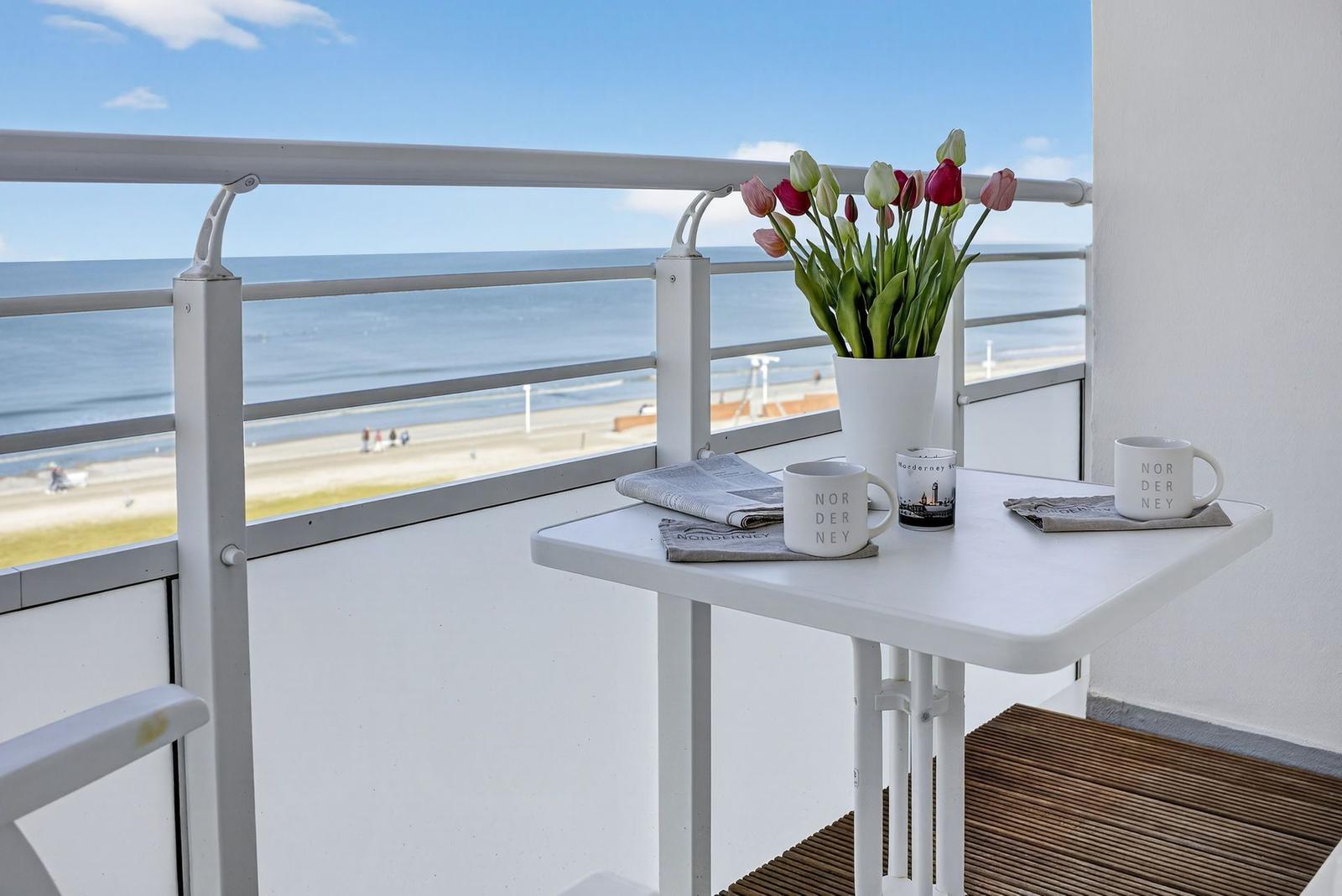 Balcony with table, flowers, and sea view. Mugs with Norderney are ready.