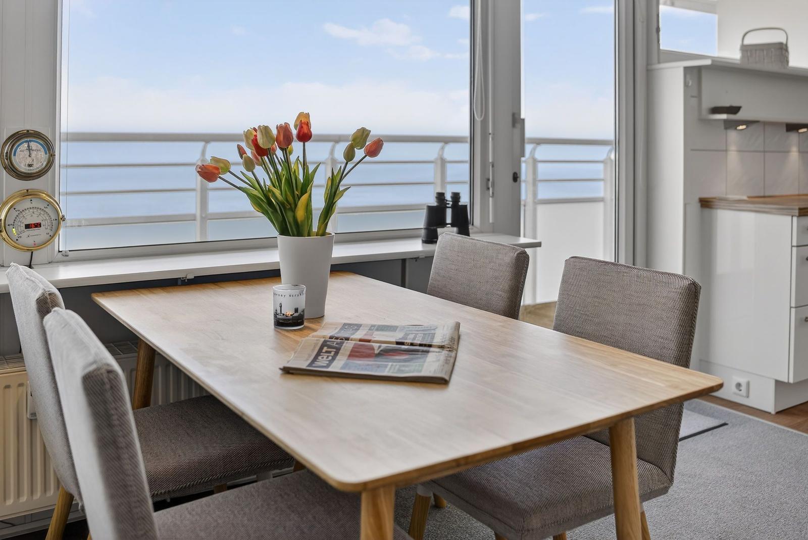 Dining table with chairs by large window with sea view. Flowers, newspaper, and thermometer are visible.