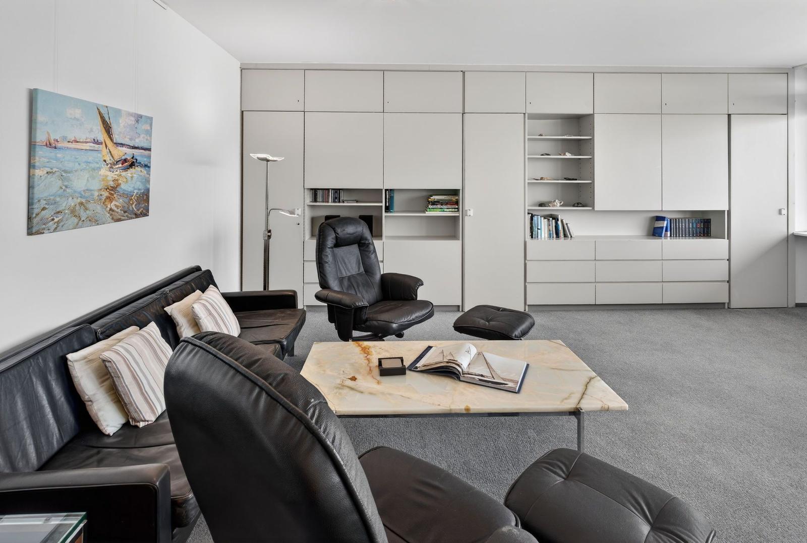 Living room with leather sofa, chairs, and marble table. Wall shelves and artwork are visible.