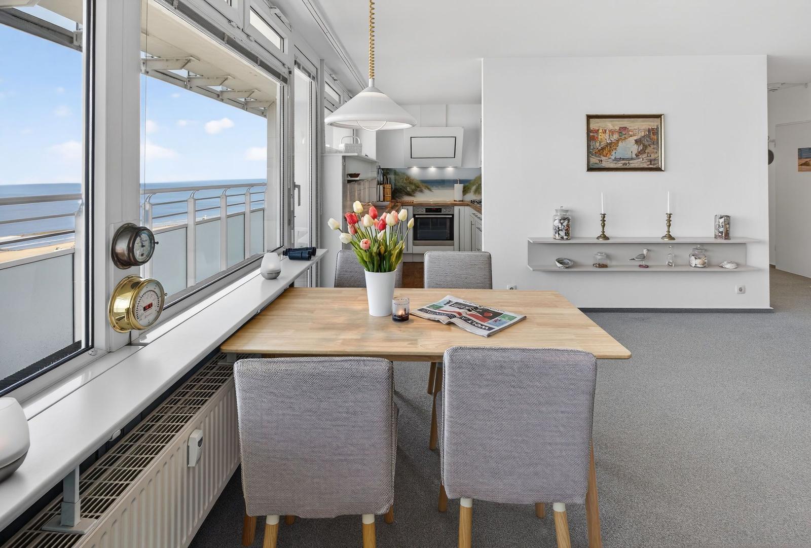 Dining area with sea view. Wooden table, chairs, flowers, and kitchen area in the background.