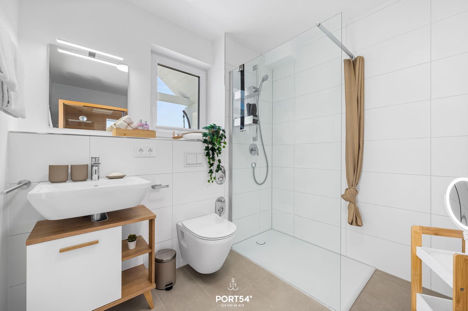 Badezimmer mit Dusche, Waschbecken und Toilette. Weiße Fliesen und Holzelemente.