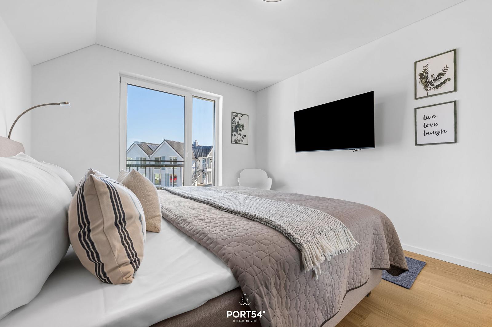 Schlafzimmer mit Bett, Fernseher und Fensterblick auf Gebäude.