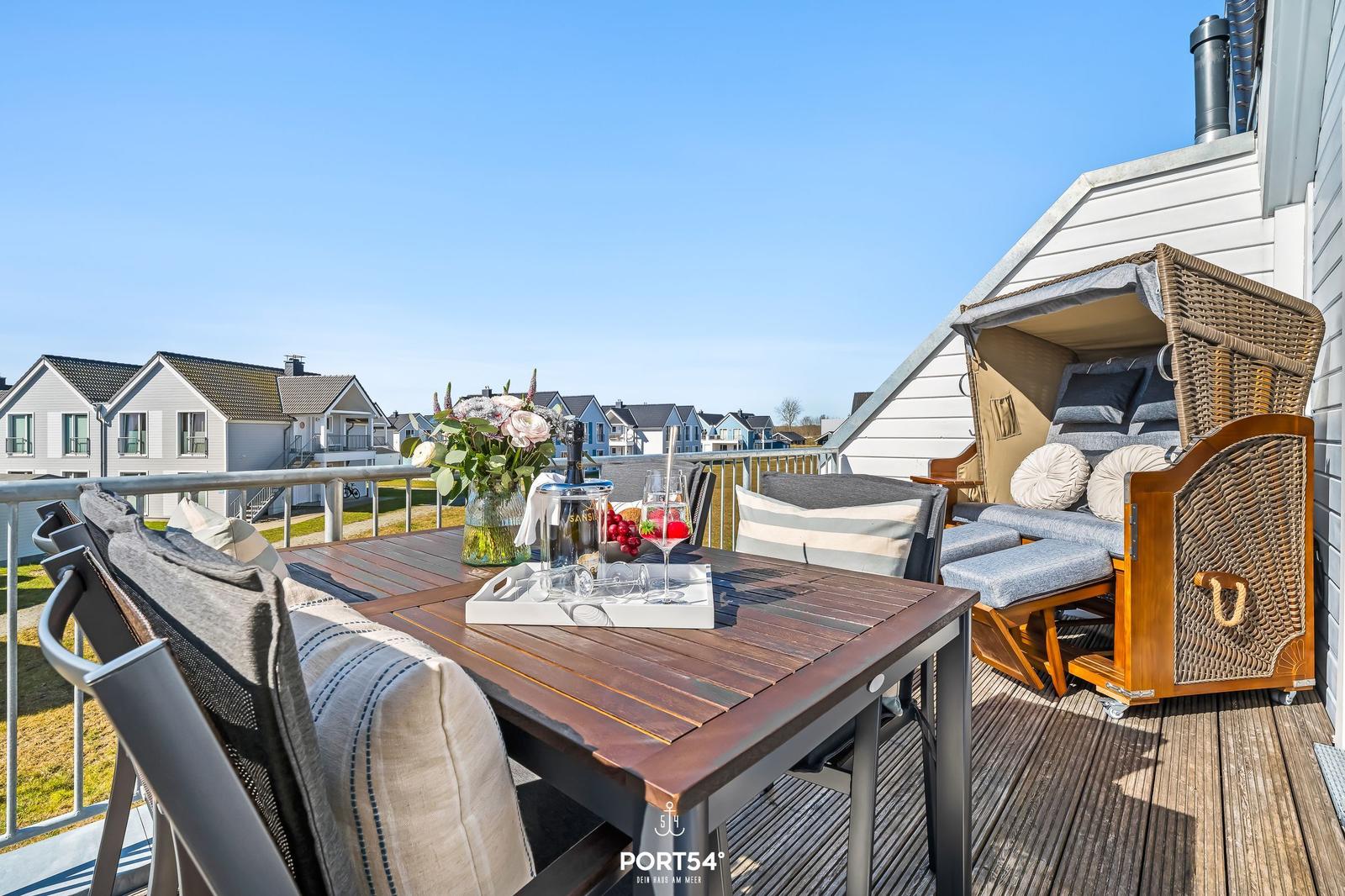 Terrasse mit Tisch, Stühlen und Strandkorb. Blick auf Wohnhäuser und blauen Himmel.