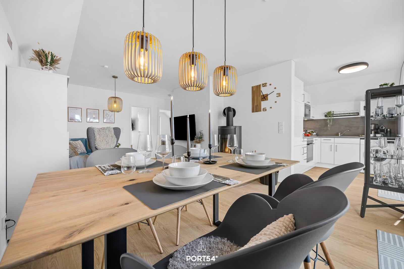 Essbereich mit Holztisch, schwarzen Stühlen und hängenden Lampen. Küche und Wohnbereich im Hintergrund.