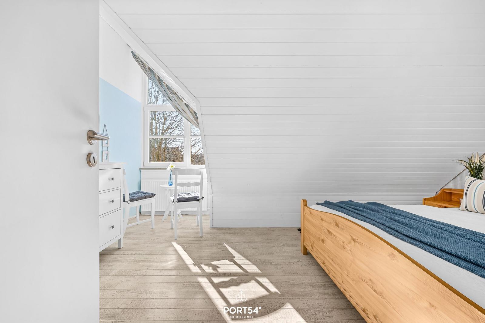 Schlafzimmer mit Holzbett, weißer Wand und Fenster mit Blick nach draußen.