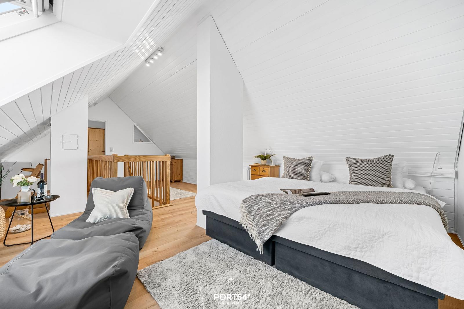 Schlafzimmer mit Doppelbett, Sitzgelegenheit und Holzboden unter schrägen weißen Wänden.