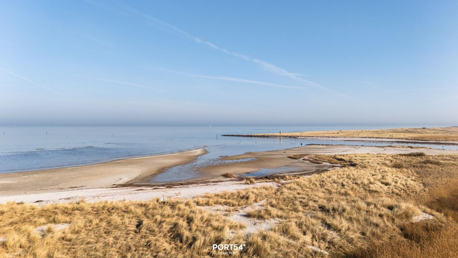Strand mit Dünen und ruhigem Meer unter blauem Himmel.