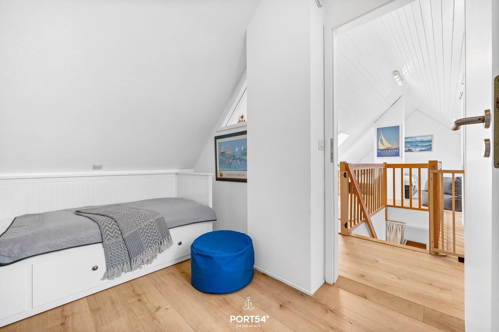 Schlafzimmer mit Dachschrägen, Bett mit Kissen und blauer Sitzhocker. Durchgang in Wohnbereich mit Holzgeländer.
