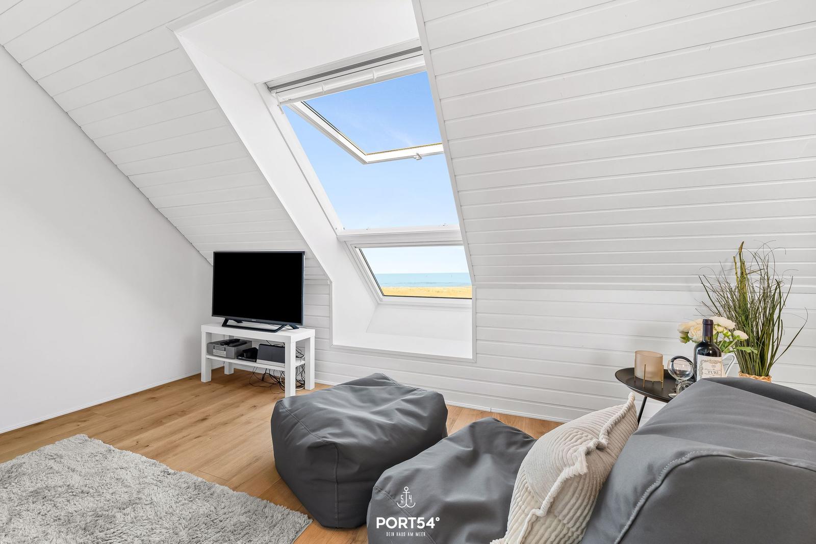 Wohnbereich mit Dachfenster, Fernseher und Sitzgelegenheiten. Blick auf Strand und Meer.