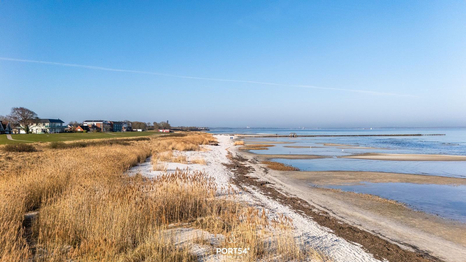 Strand mit Watt und Trockenrasen, Blick auf Häuser und Wasser.