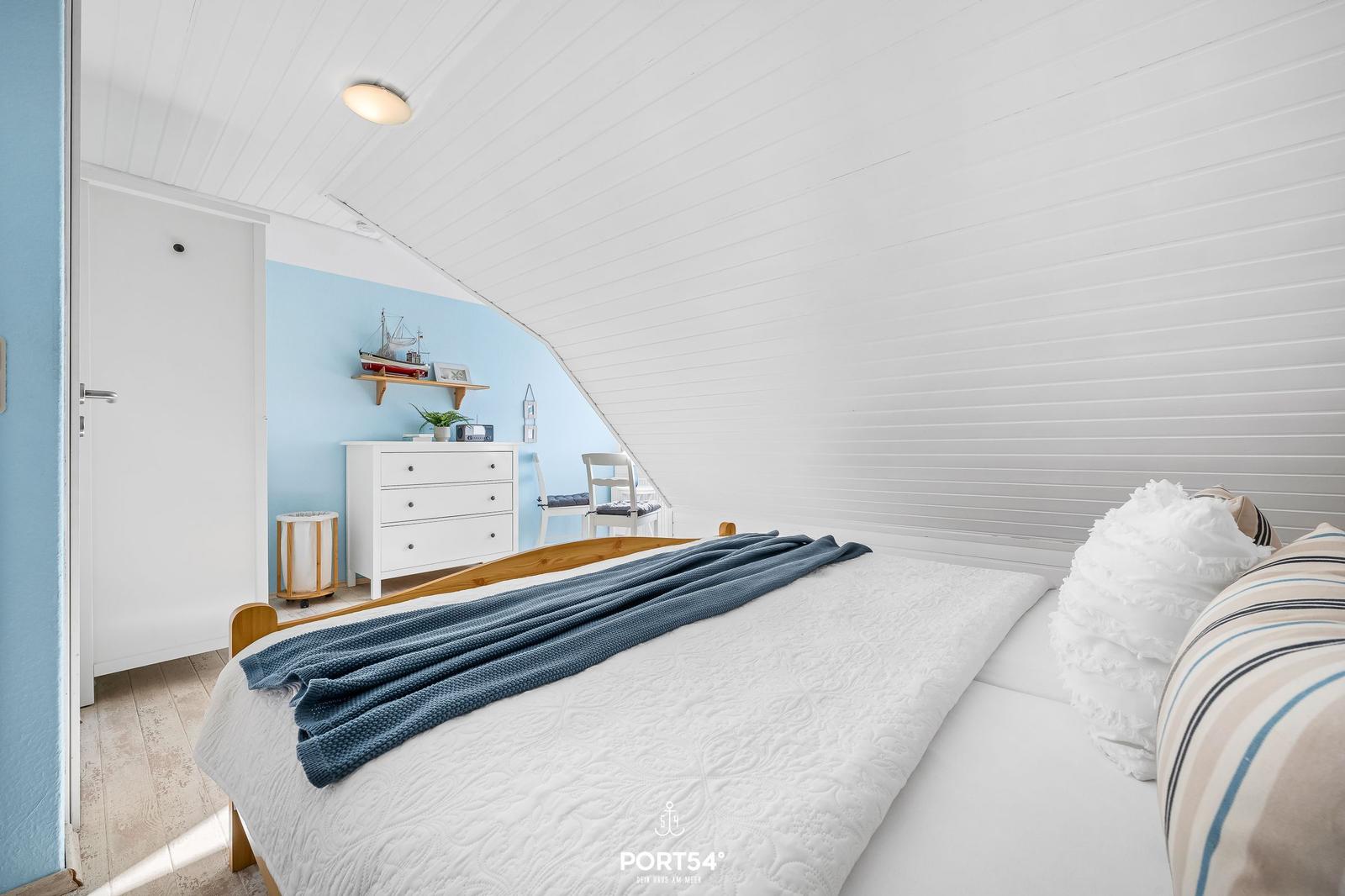 Schlafzimmer mit Dachschrägen, weißer Wand und blauer Wand. Bett, Schrank und Stuhl sind sichtbar.