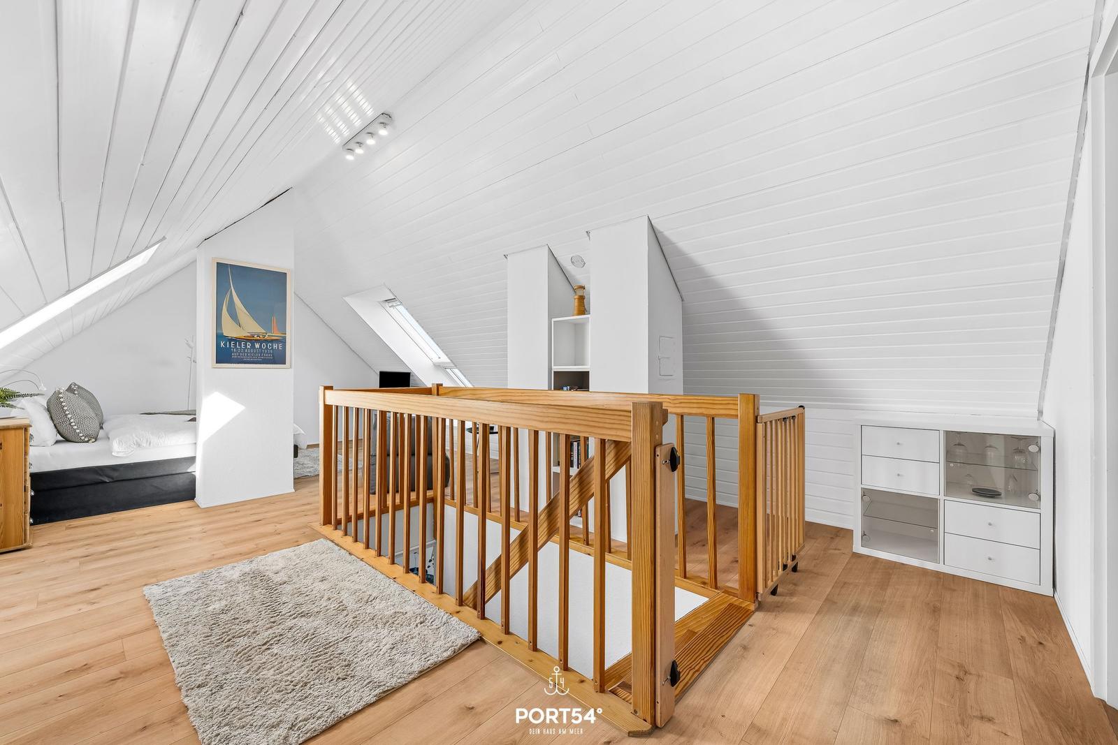 Dachgeschosszimmer mit Holzboden, weißer Decke und Holzgeländer. Bett, Schrank und Teppich sichtbar.