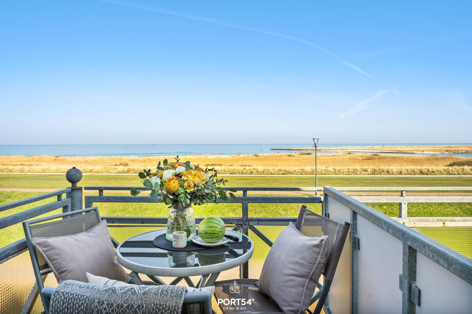 Balcony with table, chairs, and flowers overlooking sea and grass.