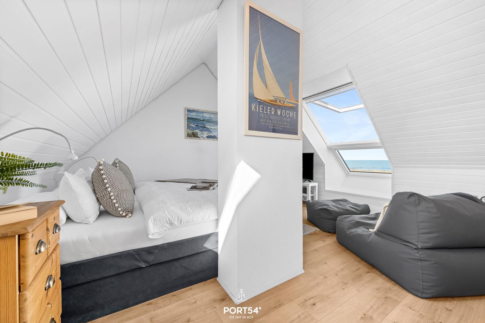 Schlafzimmer mit Dachfenster und Blick aufs Meer. Bett, Kleiderschrank und Sitzgelegenheiten.