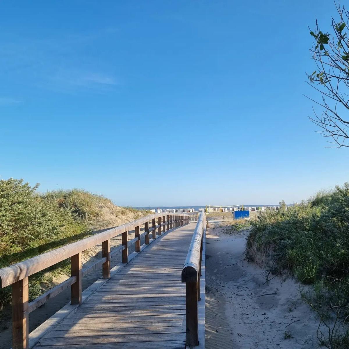 Holzweg führt durch Dünen zum Strand. Blauer Himmel und Strandkörbe sind sichtbar.