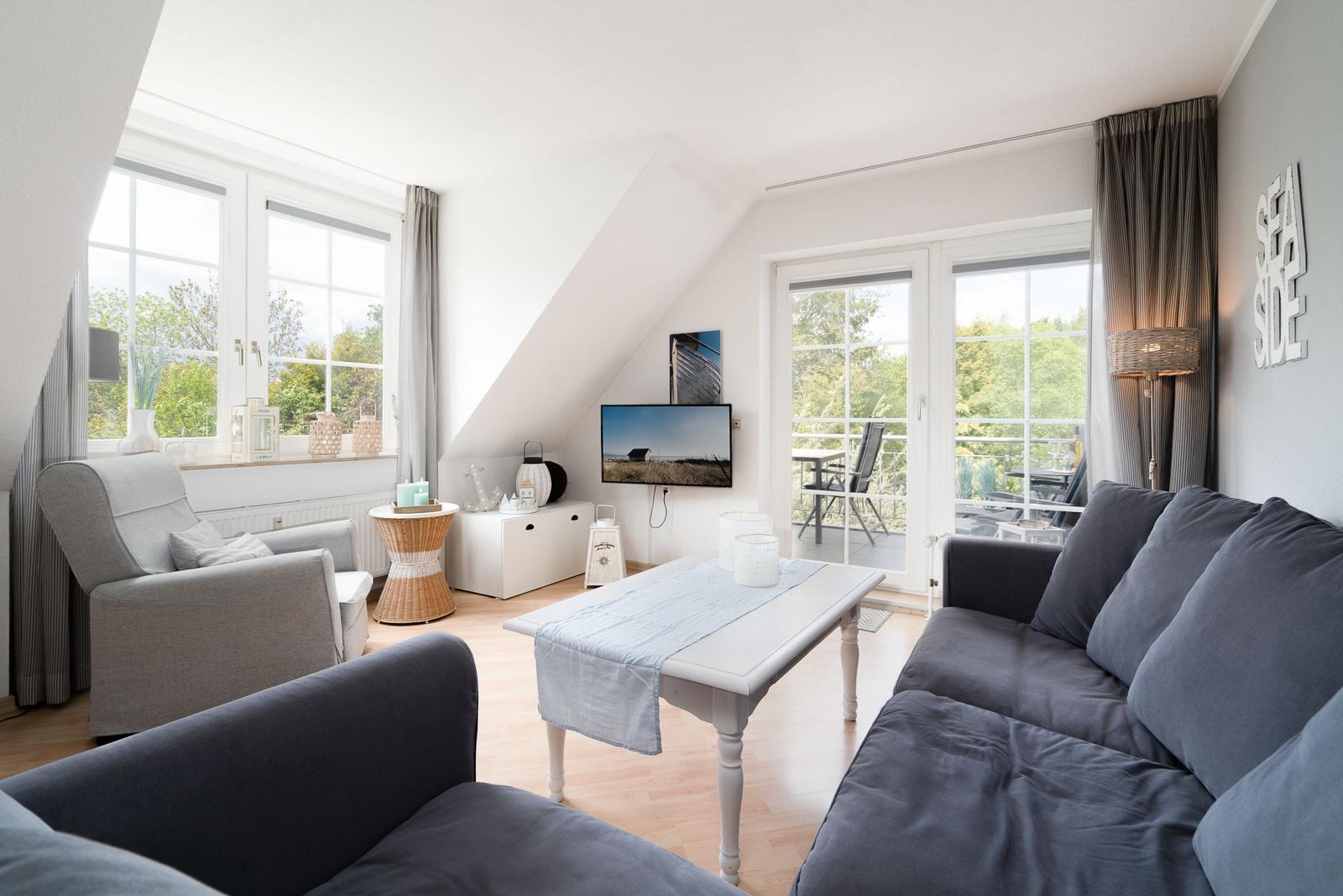 Wohnzimmer mit Sofa, Sessel und TV. Balkon mit Sicht auf Grün.