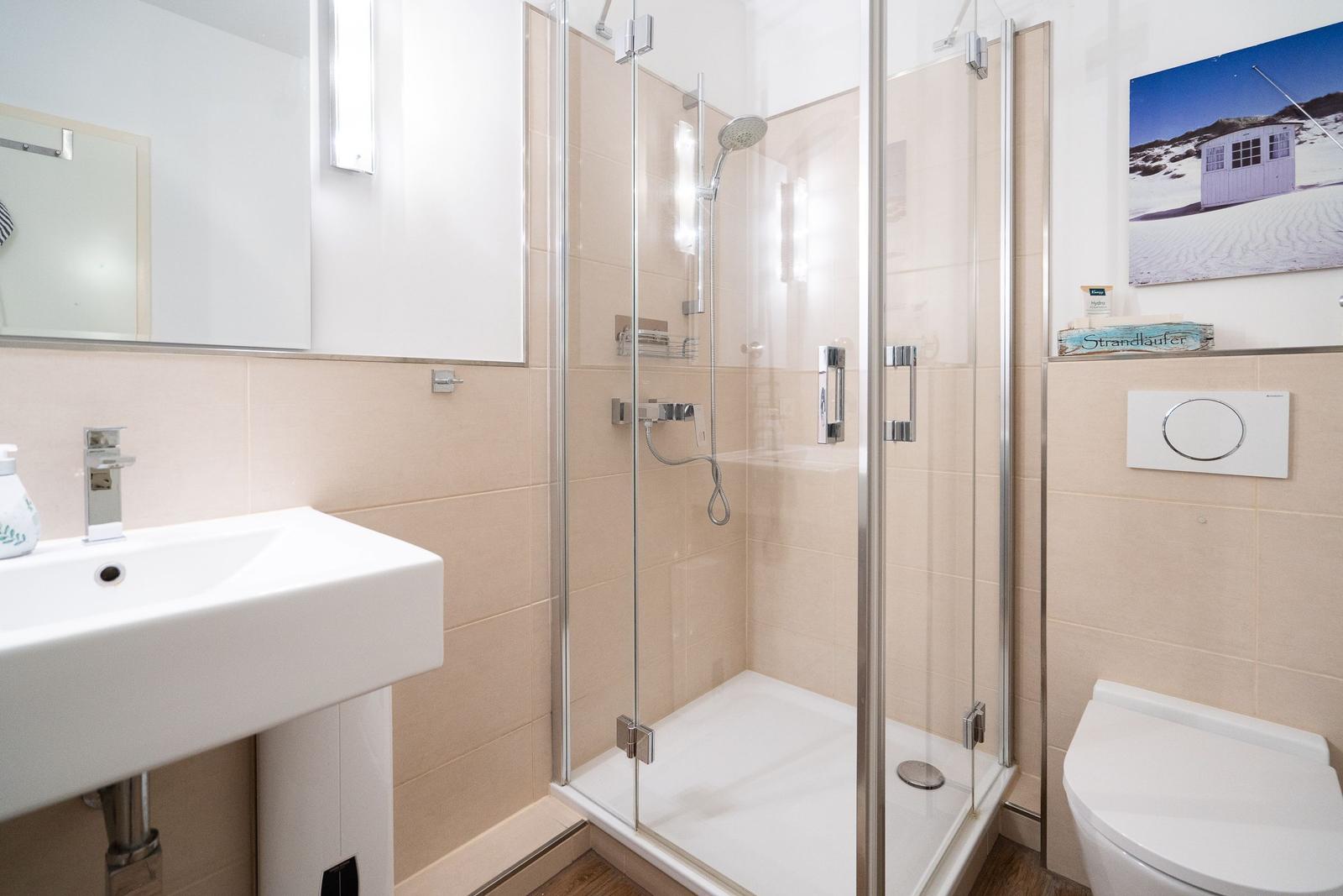 Badezimmer mit Dusche, Waschbecken und Toilette. Wandbild zeigt Strandhaus.