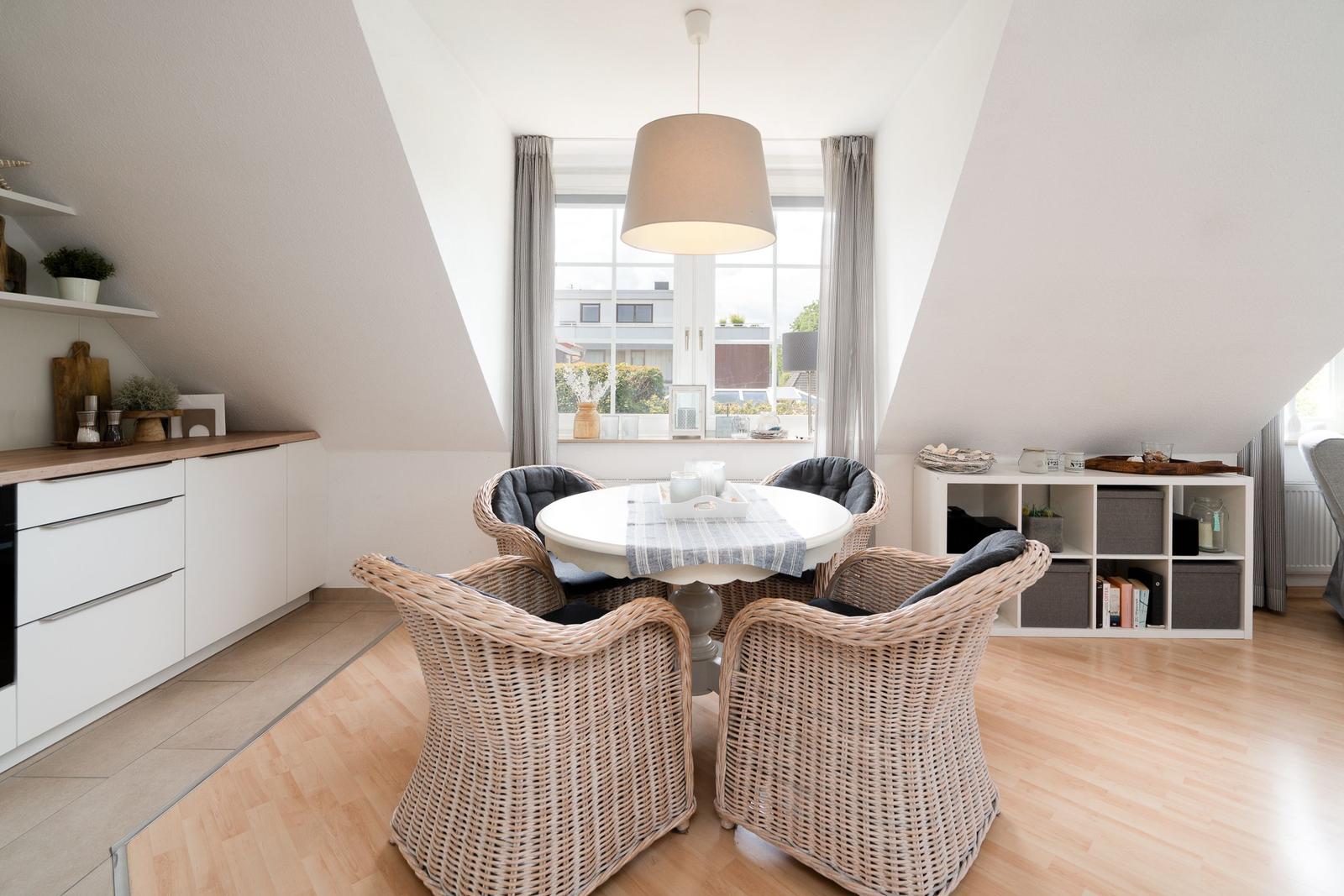 Esszimmer mit runden Tisch, vier Stühlen und großem Fenster. Weiße Küchenzeile und Holzboden.