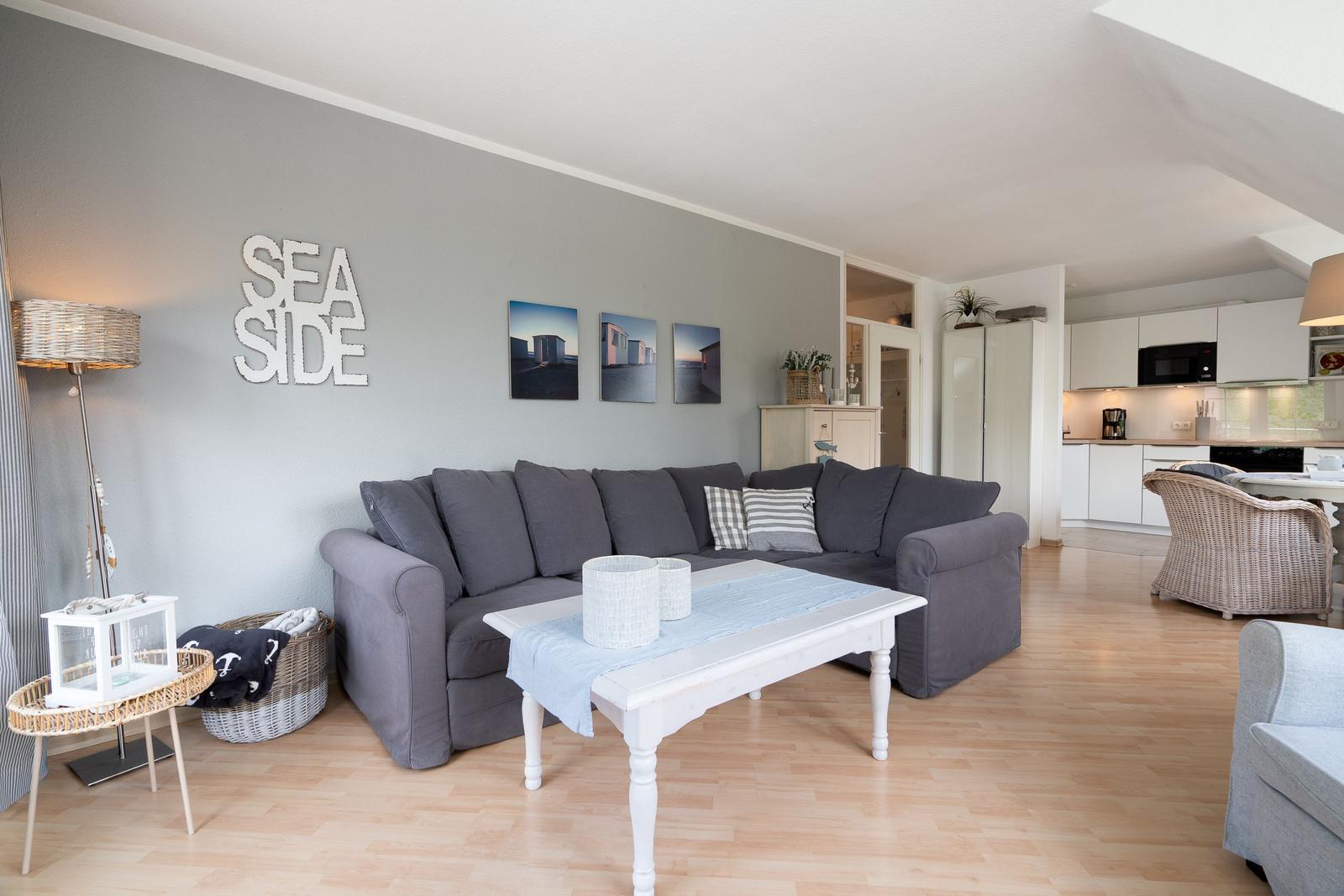 Wohnzimmer mit grauem Sofa, weißem Tisch und Küchenbereich im Hintergrund.