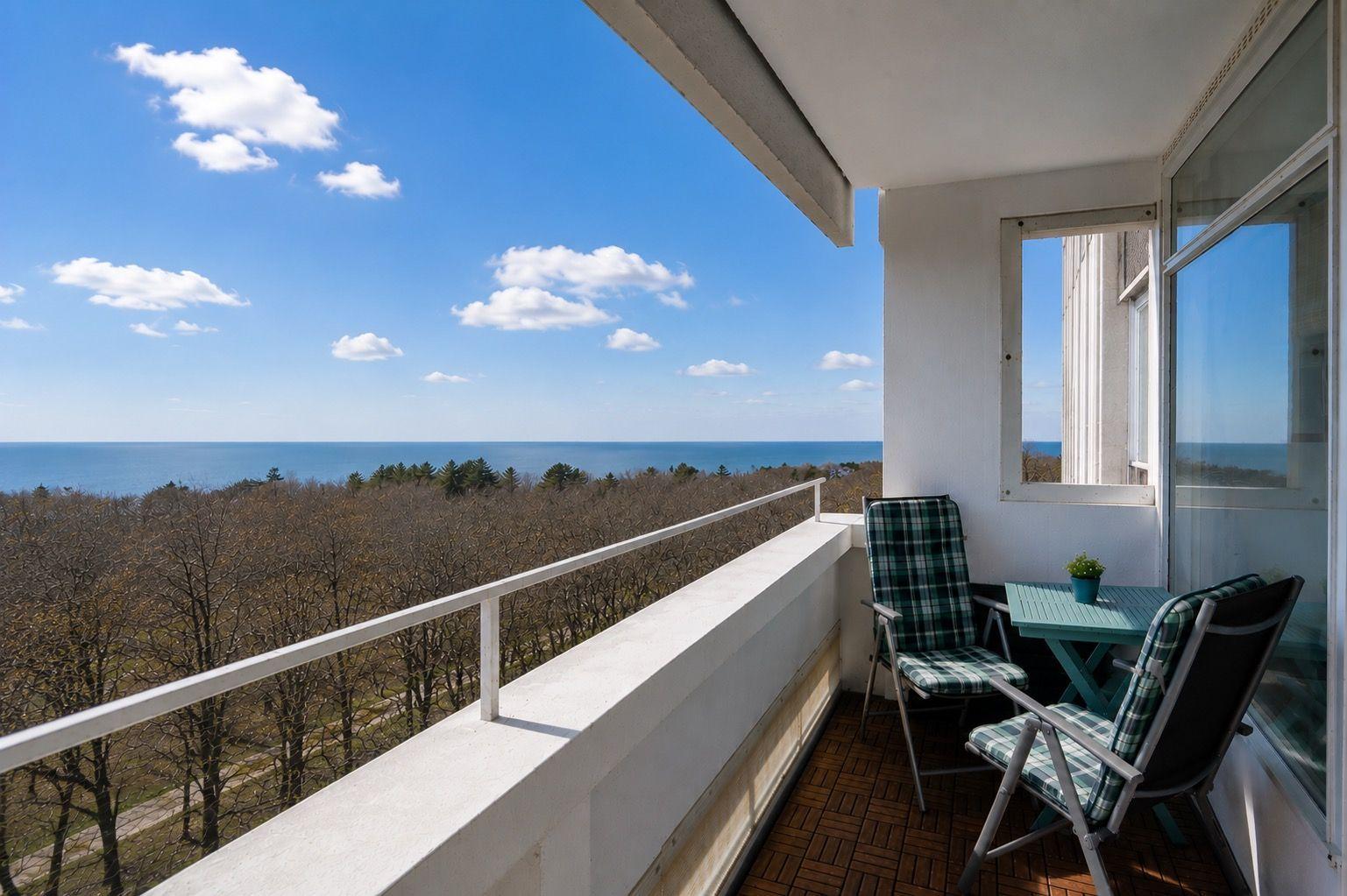 Balcony with table and chairs overlooking sea and forest under blue sky.