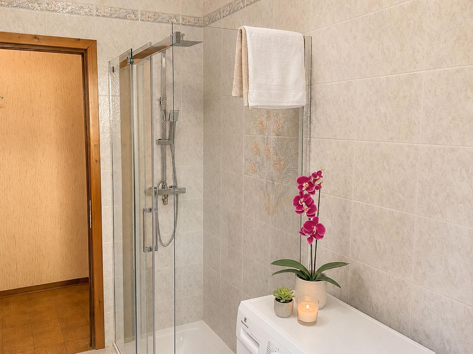 Shower with glass enclosure, towel rack, and plants on washing machine.