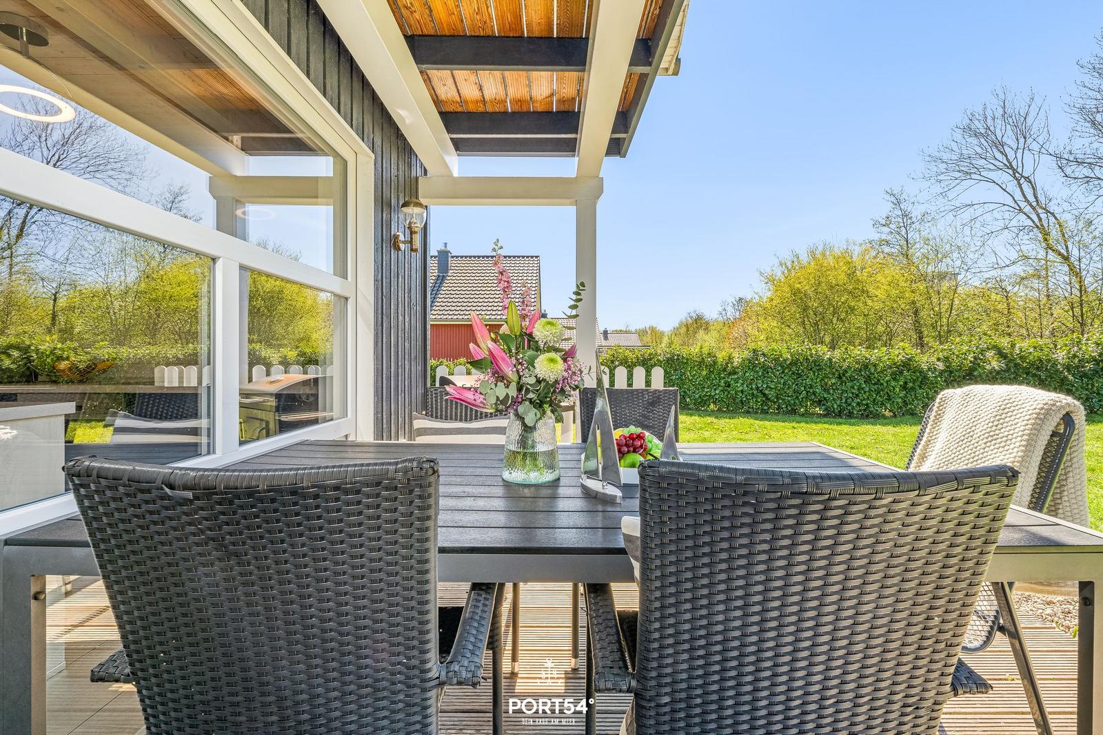 Terrasse mit Tisch und Stühlen, Blumen und Obst. Blick auf grünen Garten und Himmel.