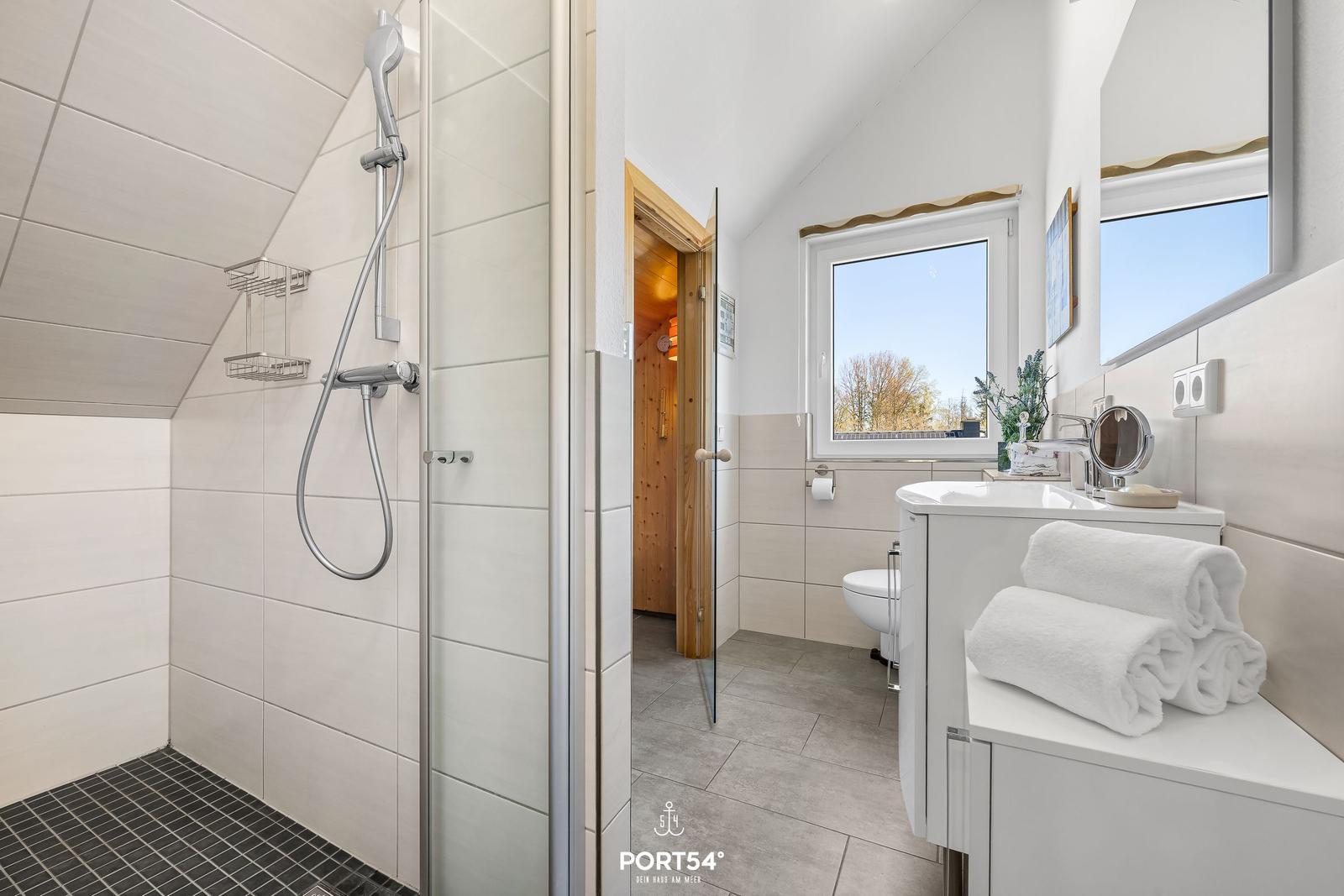 Badezimmer mit Dusche, Waschbecken und Toilette. Fenster mit Blick nach draußen.