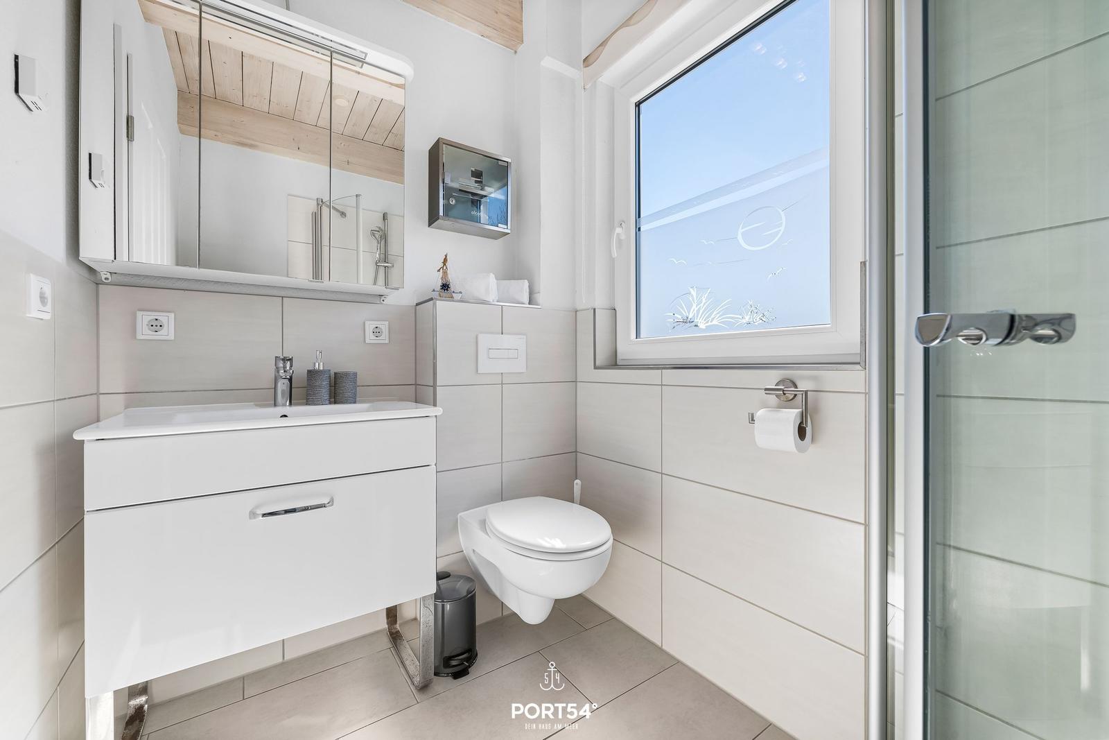 Badezimmer mit Waschbecken, Toilette und Dusche. Fenster mit Motiv und Spiegelschrank.