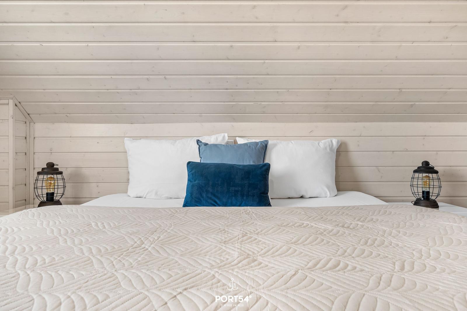 Ein Bett mit weißer Bettwäsche und blauen Kissen vor einer hellen Holzwand.