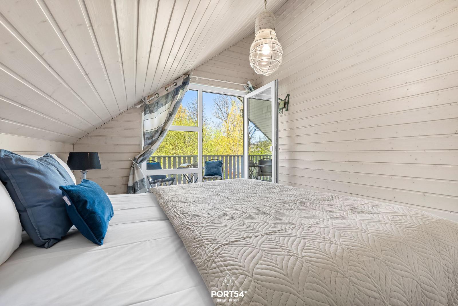 Schlafzimmer mit Bett, Balkon und Holzwand. Deckenleuchte und Fenster sichtbar.