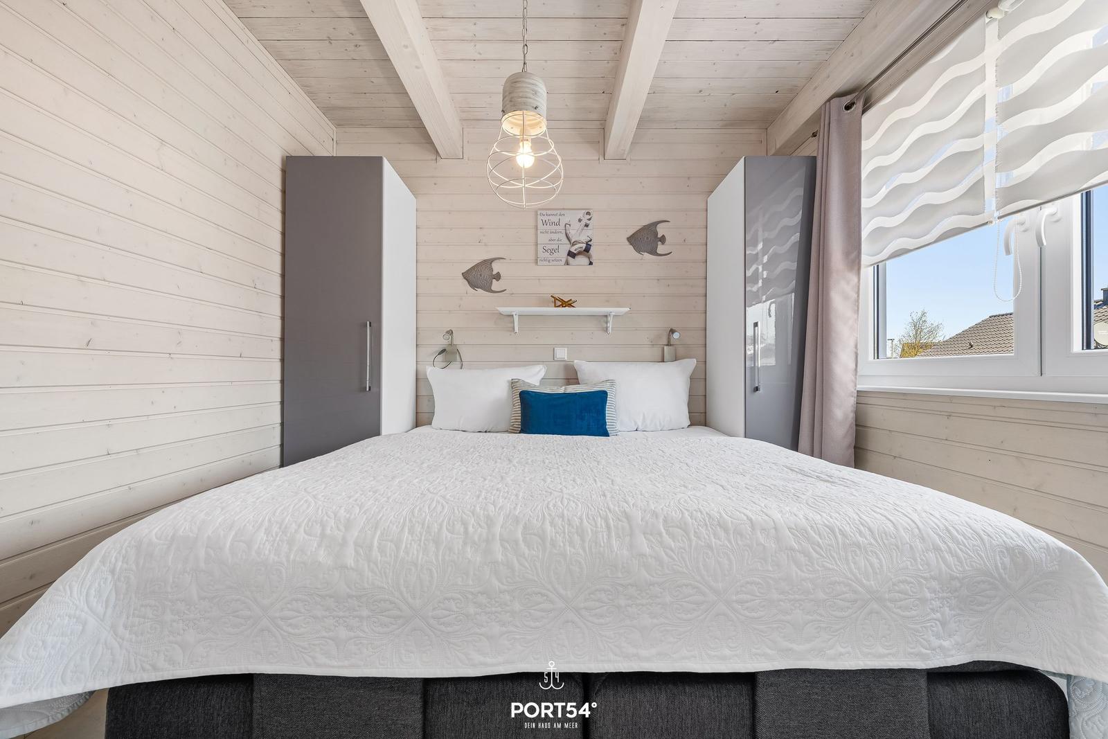 Schlafzimmer mit Doppelbett, weißen Wänden und Holzdecke. Fenster mit Rollladen und Vorhang.