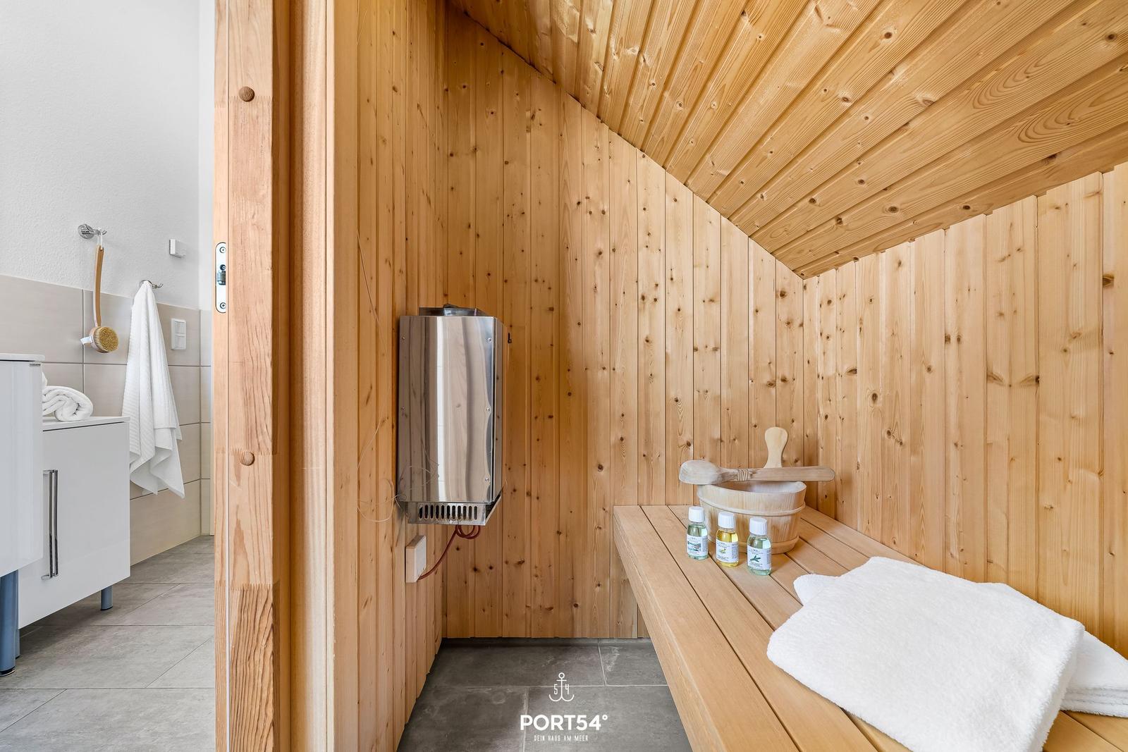 Holzsauna mit Sitzbank, Saunakorb und Dampfgenerator. Weiße Handtücher und Flaschen darauf.