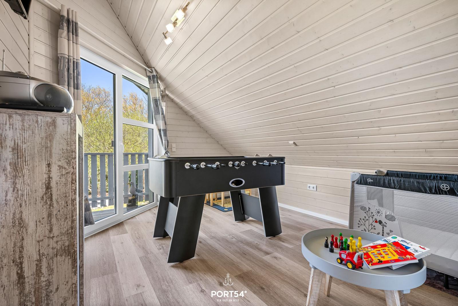 Spielzimmer mit Tischfußball, Babybett und Kinderspielzeug. Große Fenster mit Blick auf Balkon.