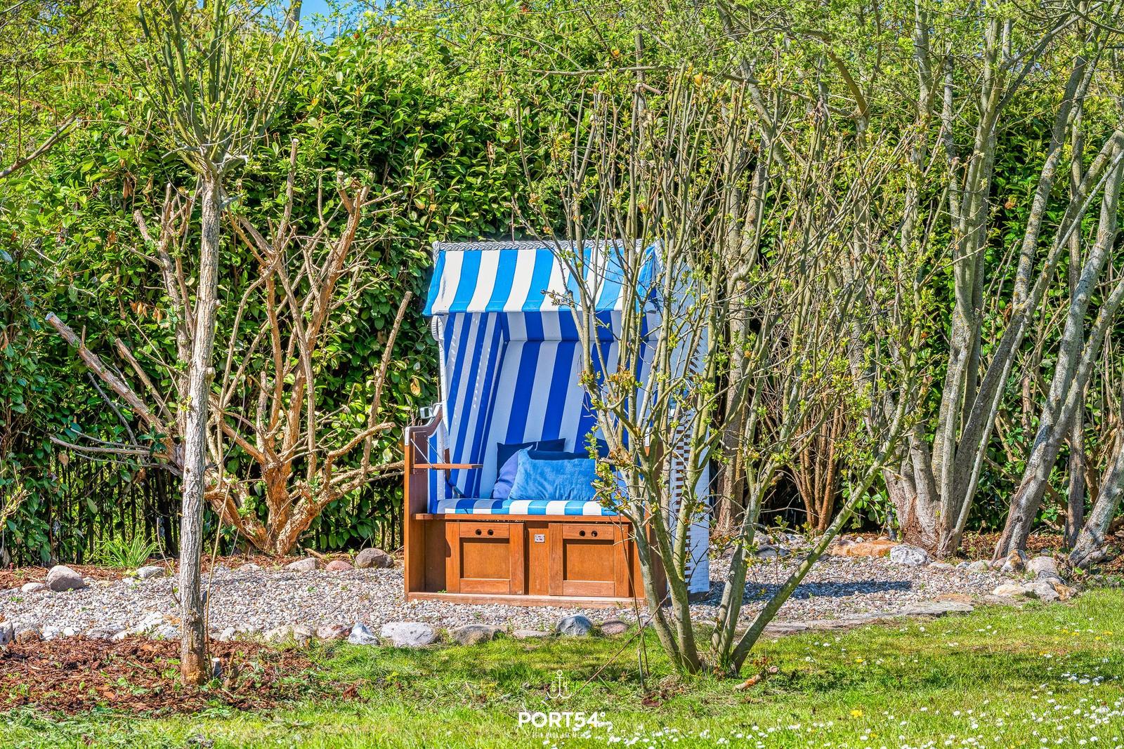 Ein blau-weiß gestreifter Strandkorb steht im Garten zwischen Bäumen und Sträuchern.