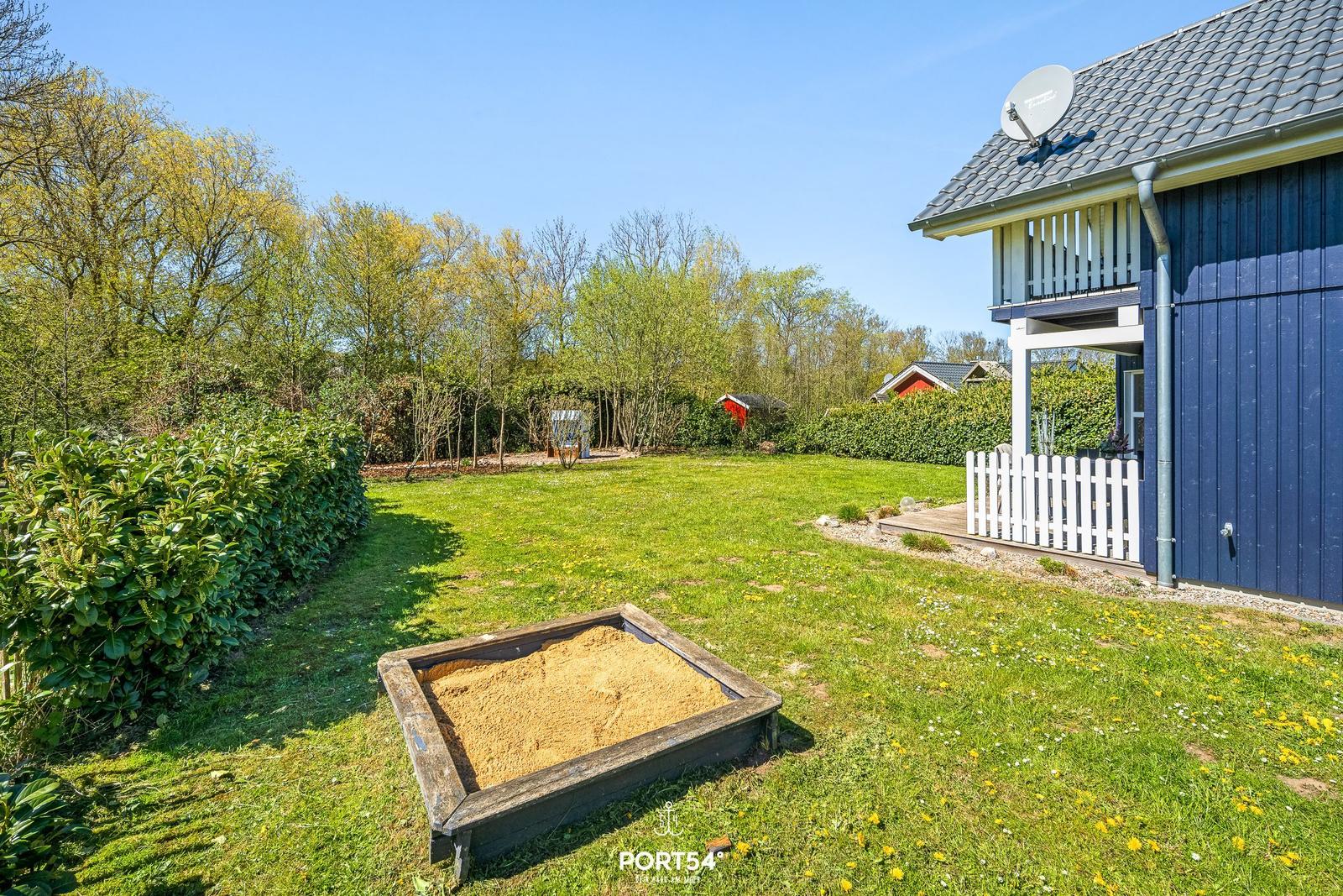 Haus mit blauer Fassade und weißem Zaun im Garten mit Sandkasten und Grünflächen.