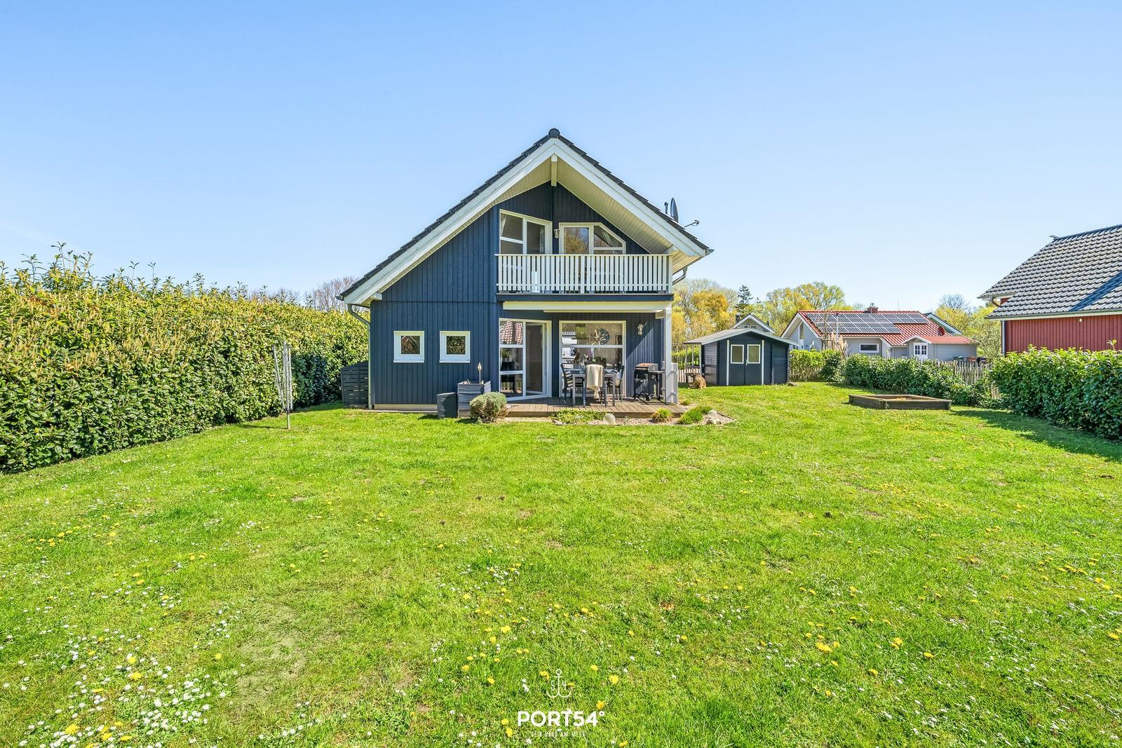 Ein zweistöckiges Haus mit Balkon und Garten. Im Hintergrund sind weitere Häuser sichtbar.