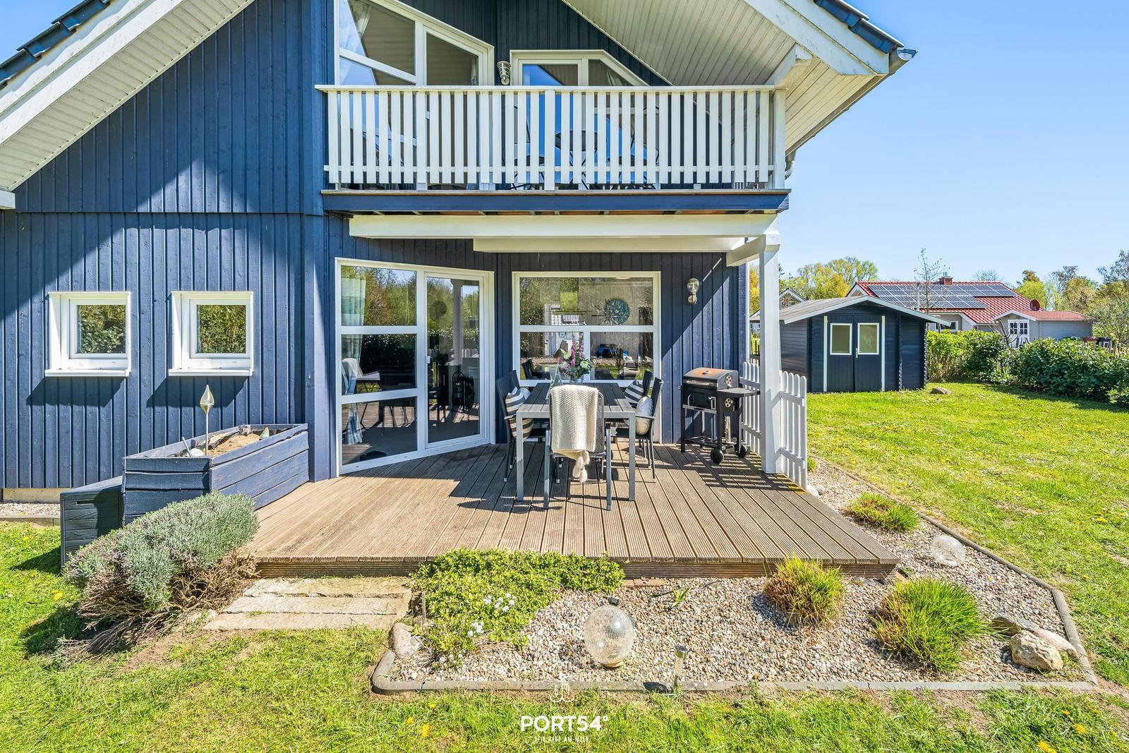 Blau gestrichenes Haus mit Terrasse, Grill und Garten. Weiße Balkonbrüstung und große Fenster.