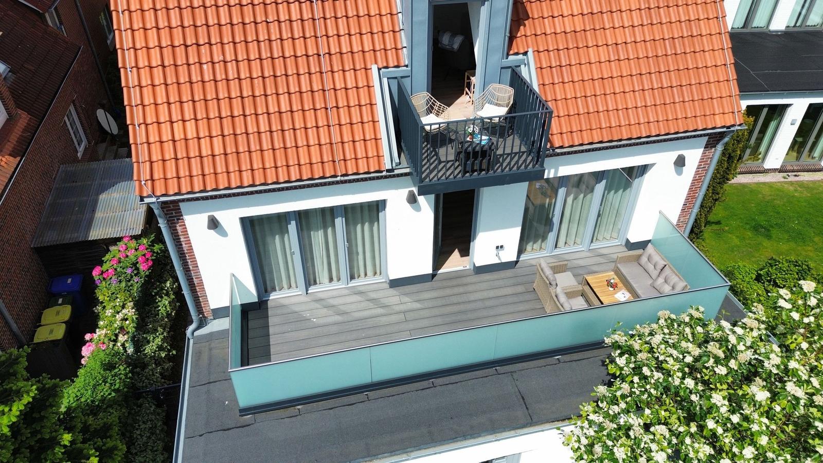 Doppelgeschossiges Haus mit Balkon und Terrasse, umgeben von Grünflächen und Blumen.