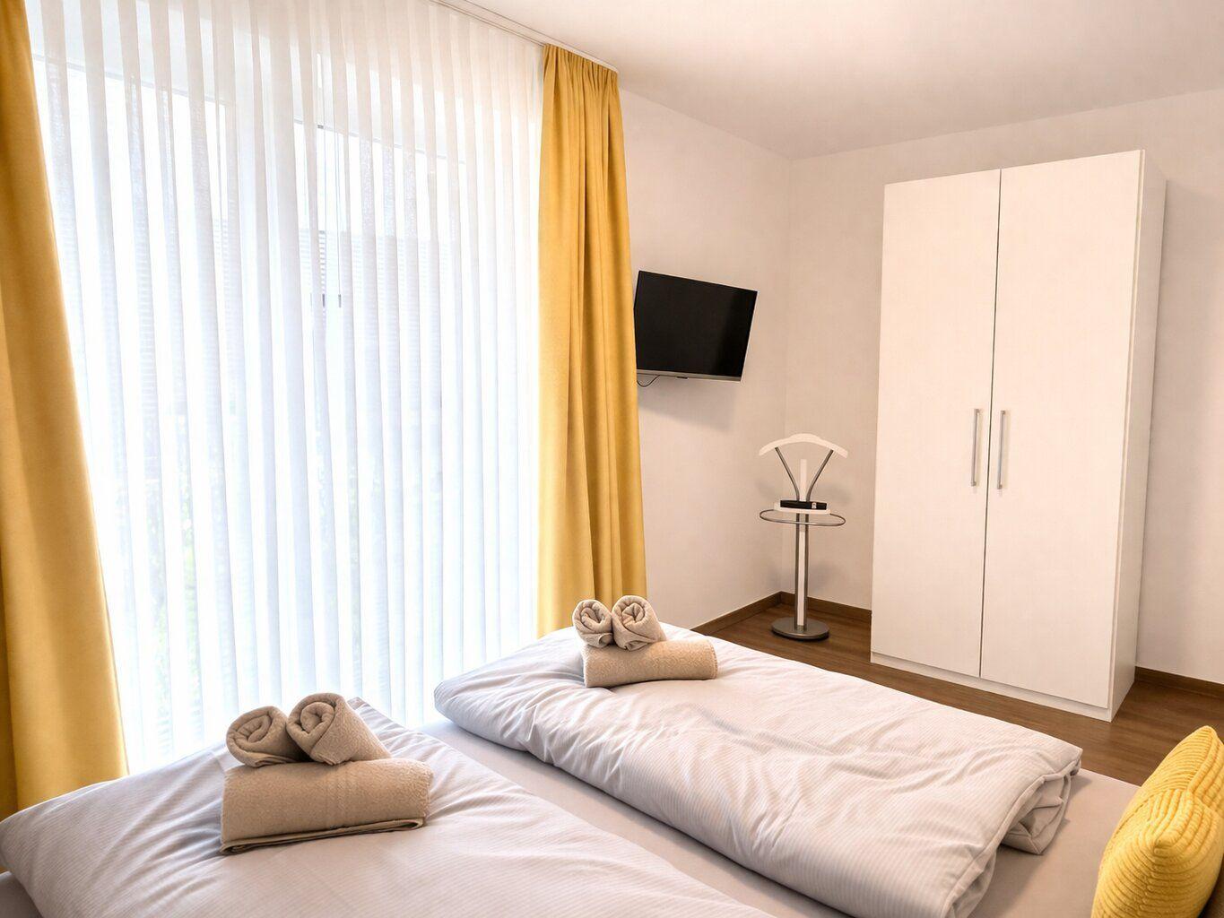 Zwei Betten mit Handtüchern, TV, Kleiderschrank und Fenster mit gelben Vorhängen.